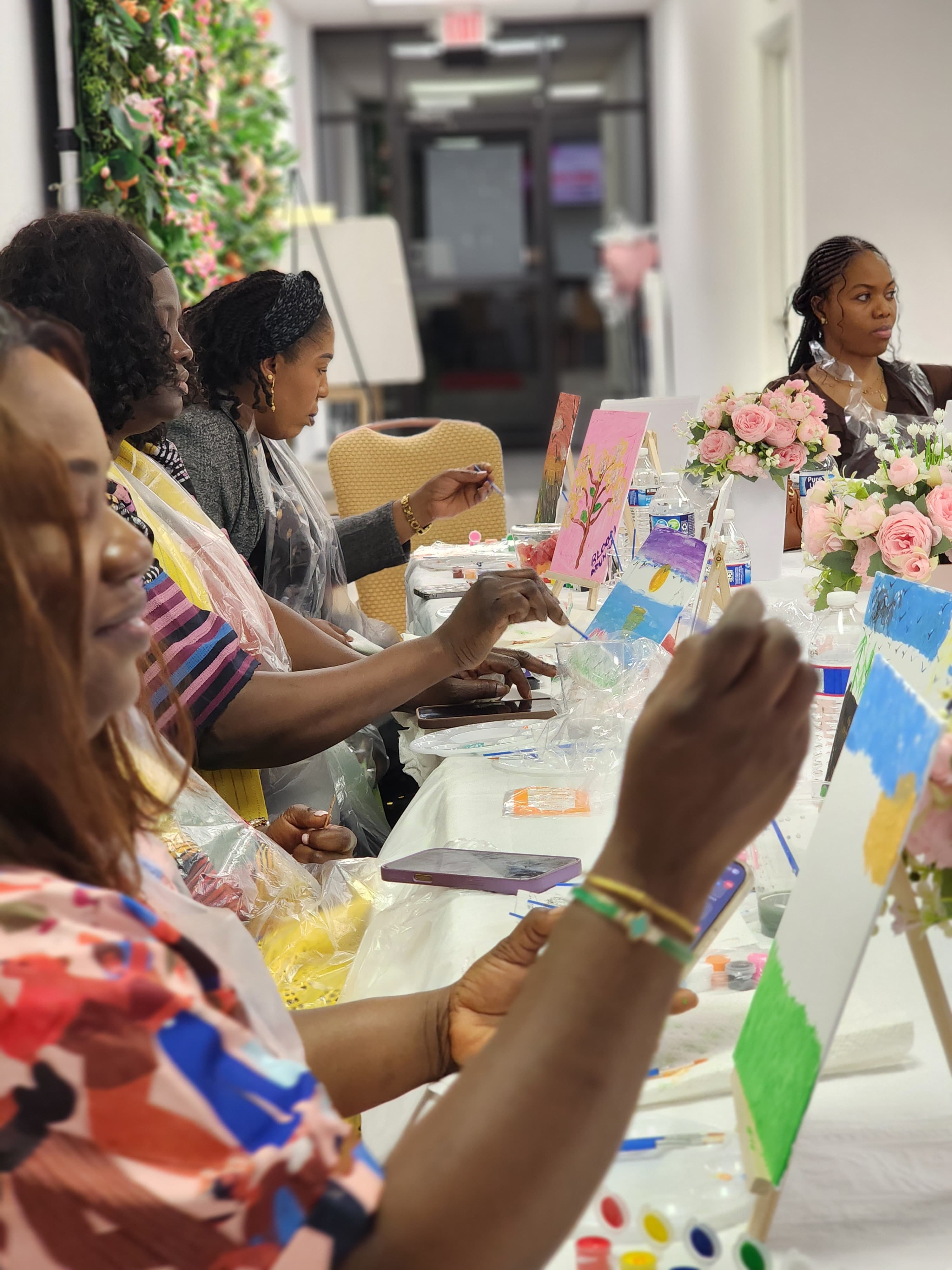 Women at a long table paint on small canvases during a social art gathering.