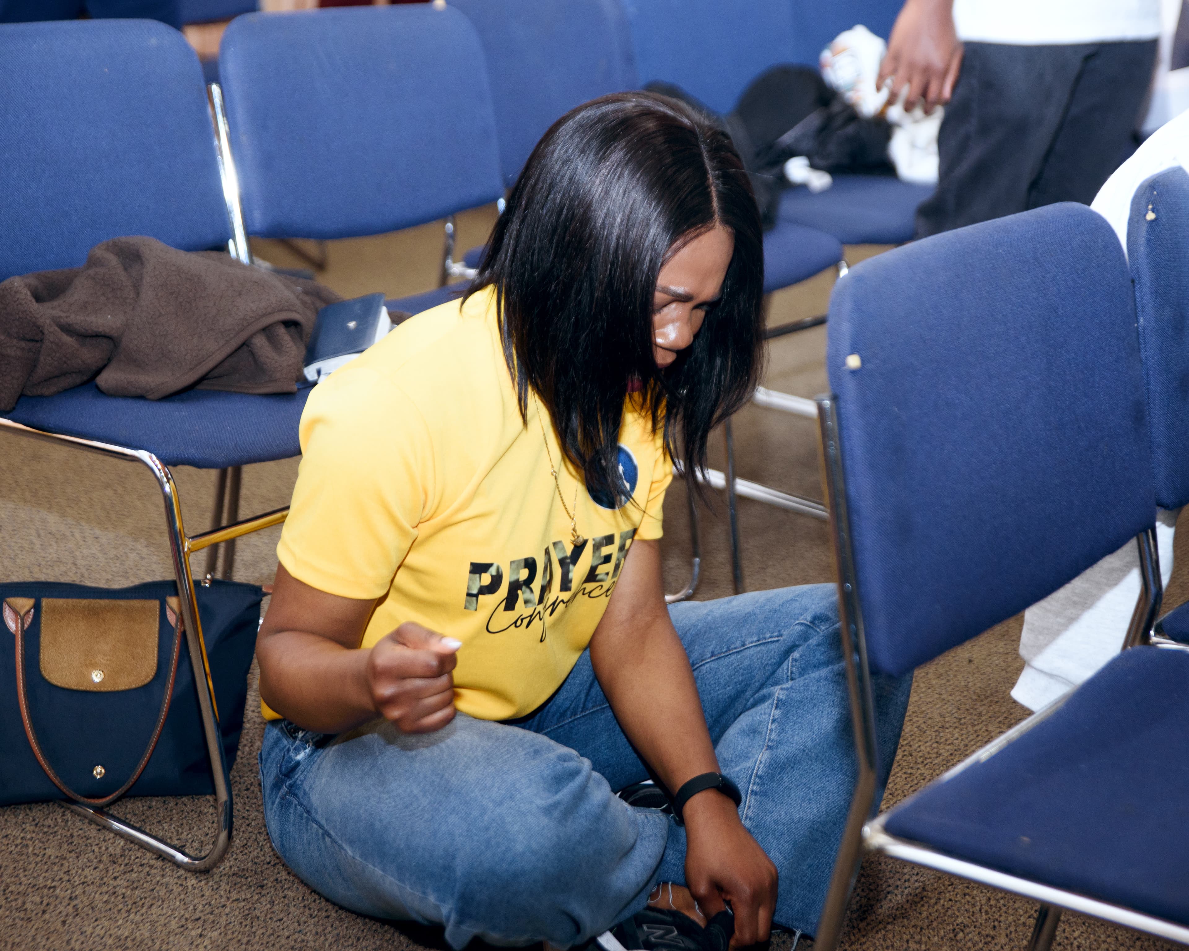 Woman in a yellow prayer conference shirt sits on floor between blue chairs, head bowed.