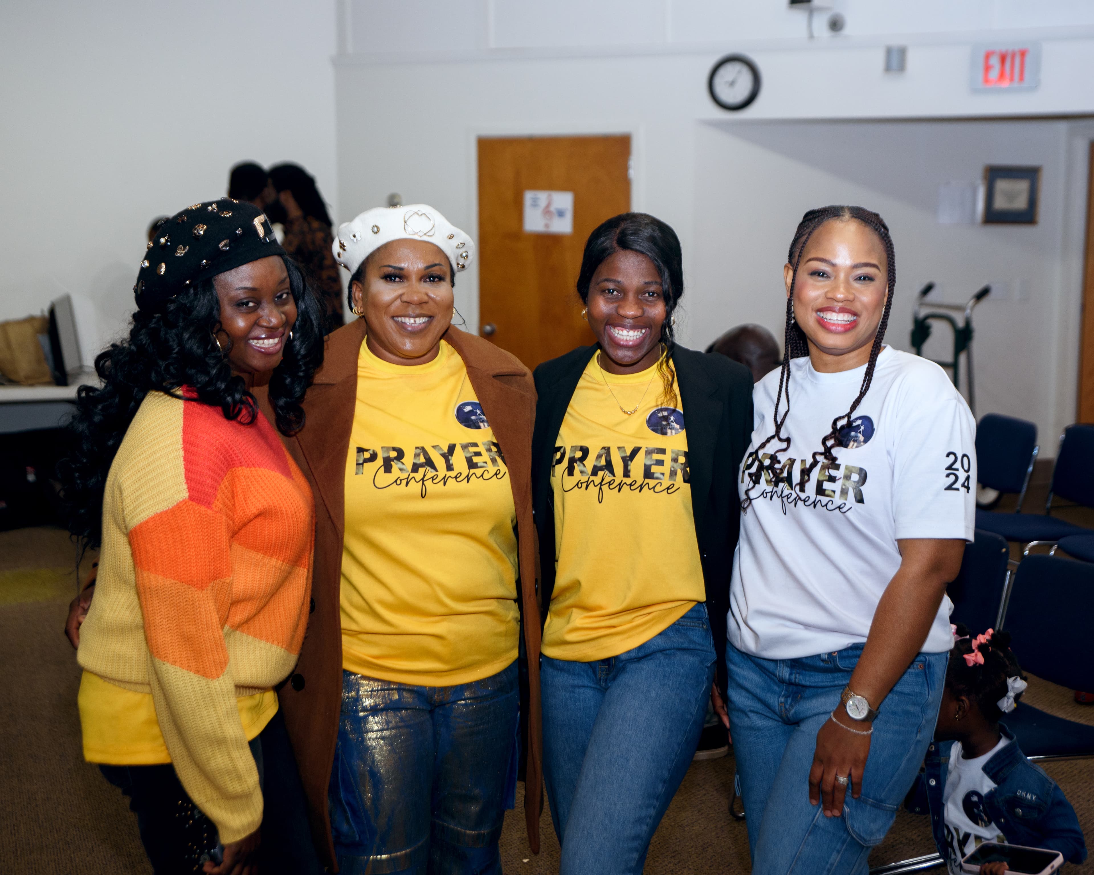 Four smiling women pose together at a prayer conference wearing yellow and white event shirts.