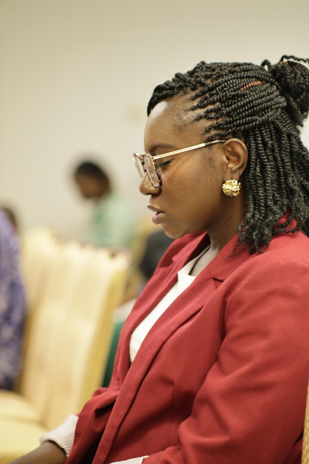 Profile of a woman with braided hair and glasses wearing a red blazer, eyes closed.
